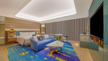A modern hotel room with a bed, a sofa, a coffee table, a TV, and a minibar.