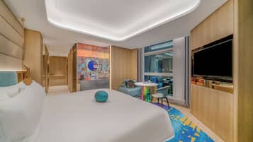 A modern hotel room with a large bed, a flat-screen TV, a seating area with a table, and a large window offering a city view.