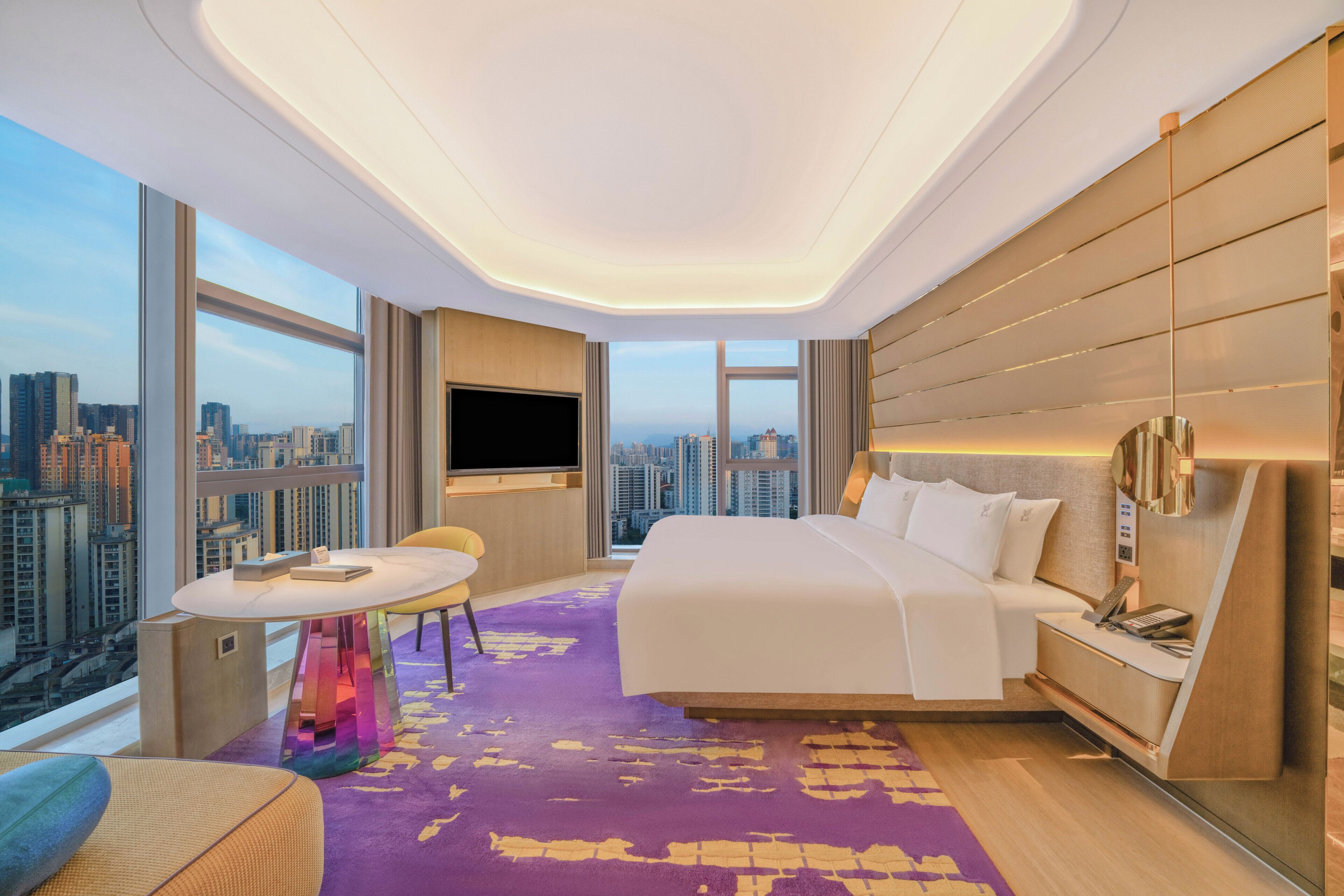 A modern hotel room with a large bed, a TV, a small table, and a view of the cityscape.