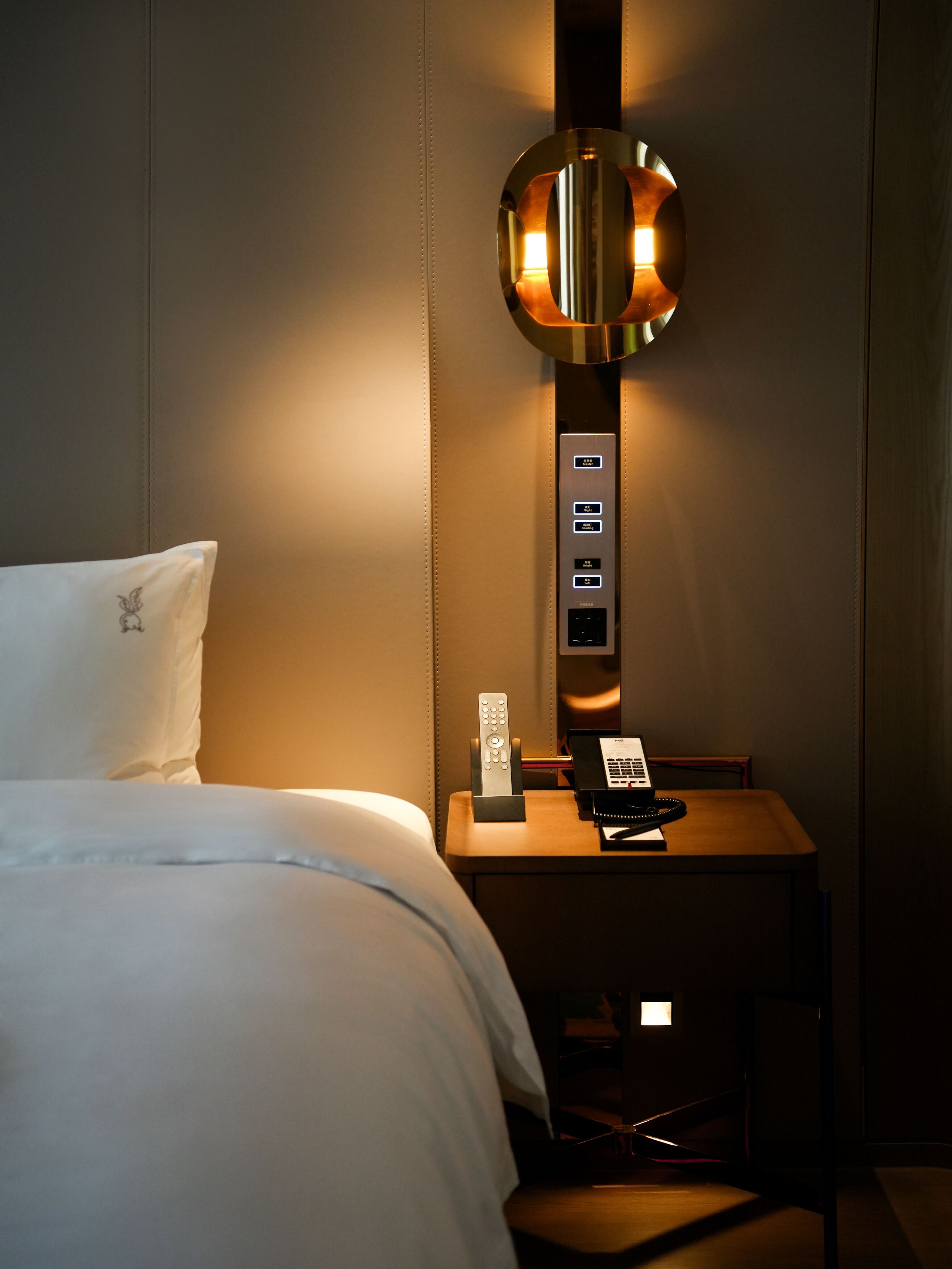 A hotel room with a bed, a bedside table with a lamp, a remote control, and a telephone.