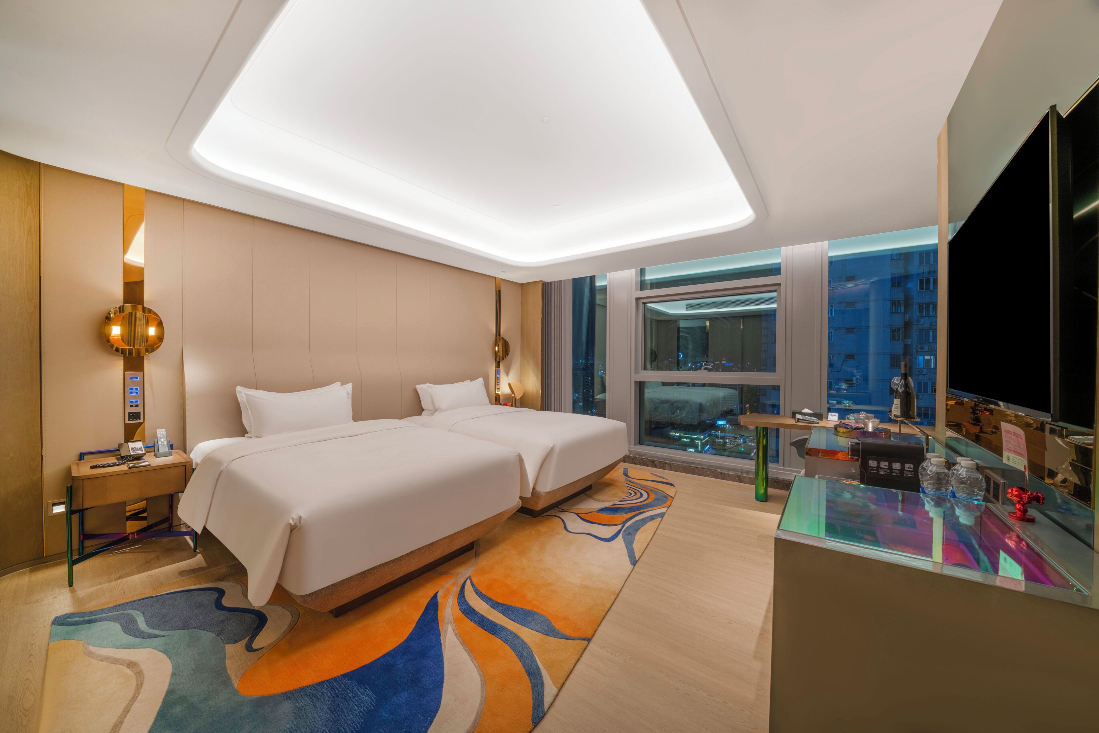 A hotel room with two beds, a desk, a desk lamp, a telephone, and a window with a city view.