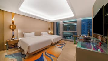 A hotel room with two beds, a desk, a desk lamp, a telephone, and a window with a city view.