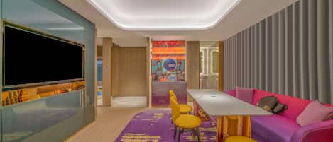 A modern hotel room with a flat-screen TV, a sofa, a dining table with chairs, and a colorful mural on the wall.