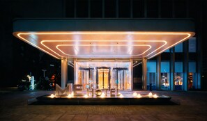A hotel entrance with a modern design, featuring a covered entrance, a fountain display, and a clear view of the interior through the glass doors.