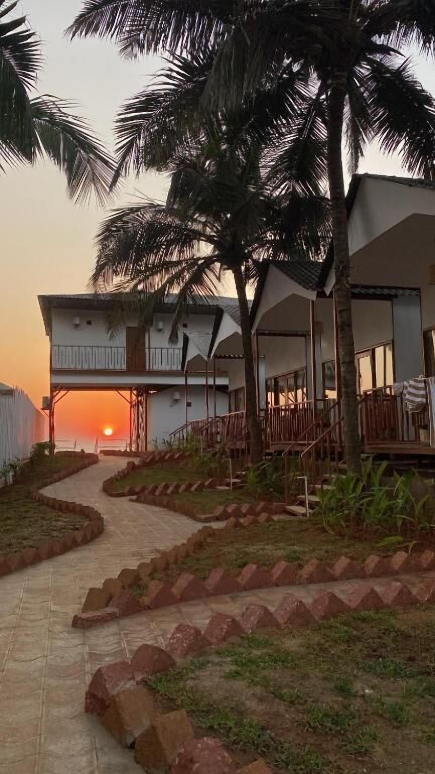 A pathway leading to a building with multiple balconies, surrounded by palm trees and a sunset in the background.