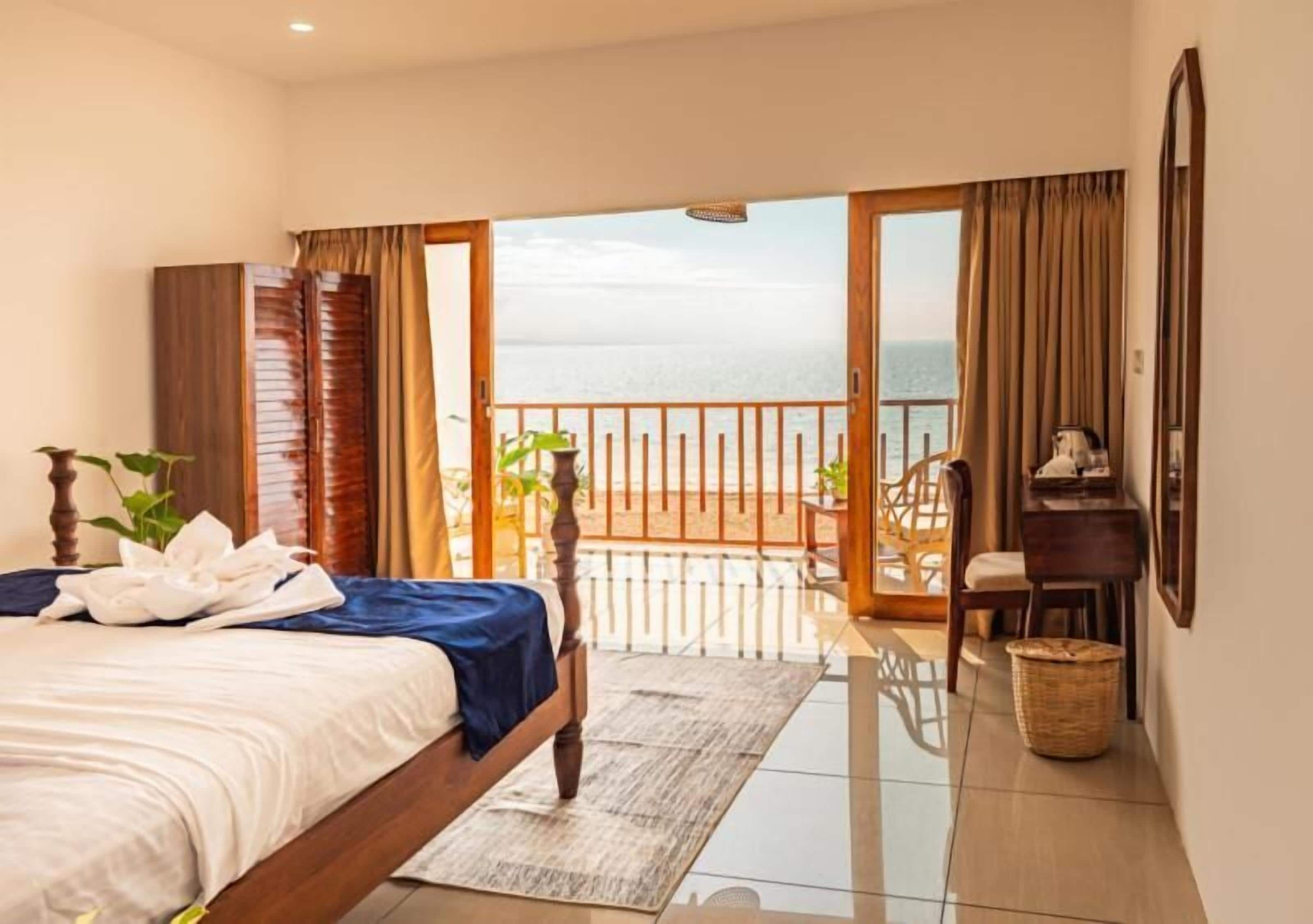 Habitación de hotel con cama, escritorio, silla, balcón con vistas al mar y ventana con cortinas.