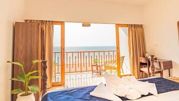Habitación de hotel con cama, escritorio, silla, planta en maceta y vista al mar a través de una puerta abierta.