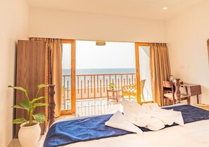 Habitación de hotel con cama, escritorio, silla, planta en maceta y vista al mar a través de una puerta abierta.
