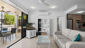 Ein modernes Wohnzimmer mit einer Couch, einem Couchtisch und einem Fernseher. Es gibt einen Balkon mit Tisch und Stühlen, sowie eine Küchenzeile mit Geräte.