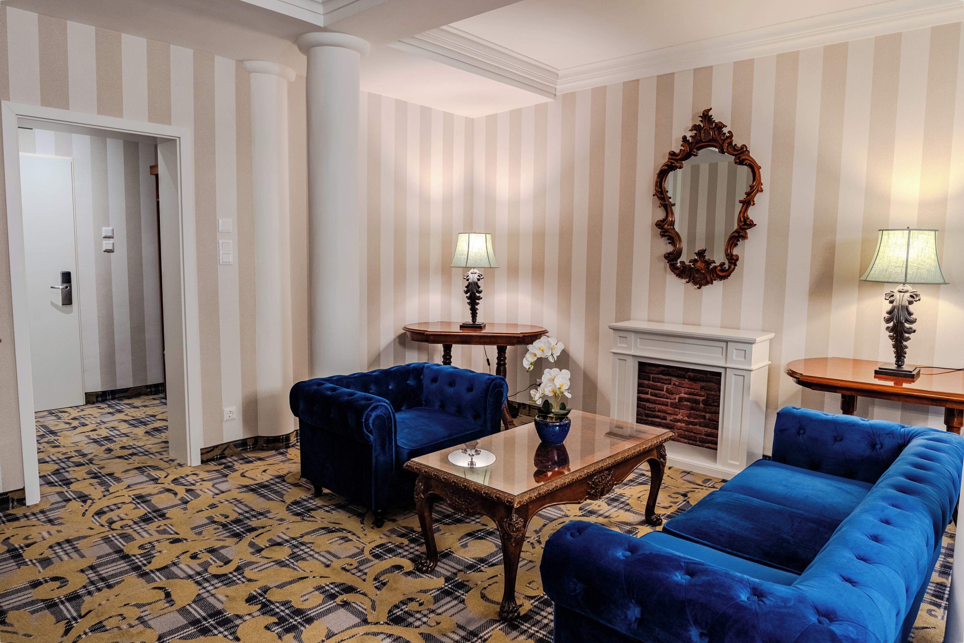 A hotel room with striped wallpaper, a fireplace, two blue sofas, a small table with a vase of flowers, and a mirror on the wall.