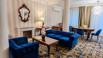 A hotel room with a blue sofa set, a wooden coffee table, a fireplace, a dining area with a round table, and a large chandelier.
