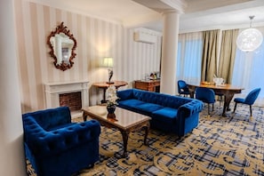 A hotel room with a blue sofa set, a wooden coffee table, a fireplace, a dining area with a round table, and a large chandelier. - Pannonia 1893 Prémium (Sopron)