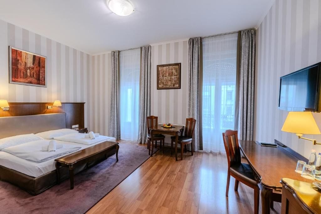 A hotel room with a bed, a desk, a chair, a television, and a painting on the wall.