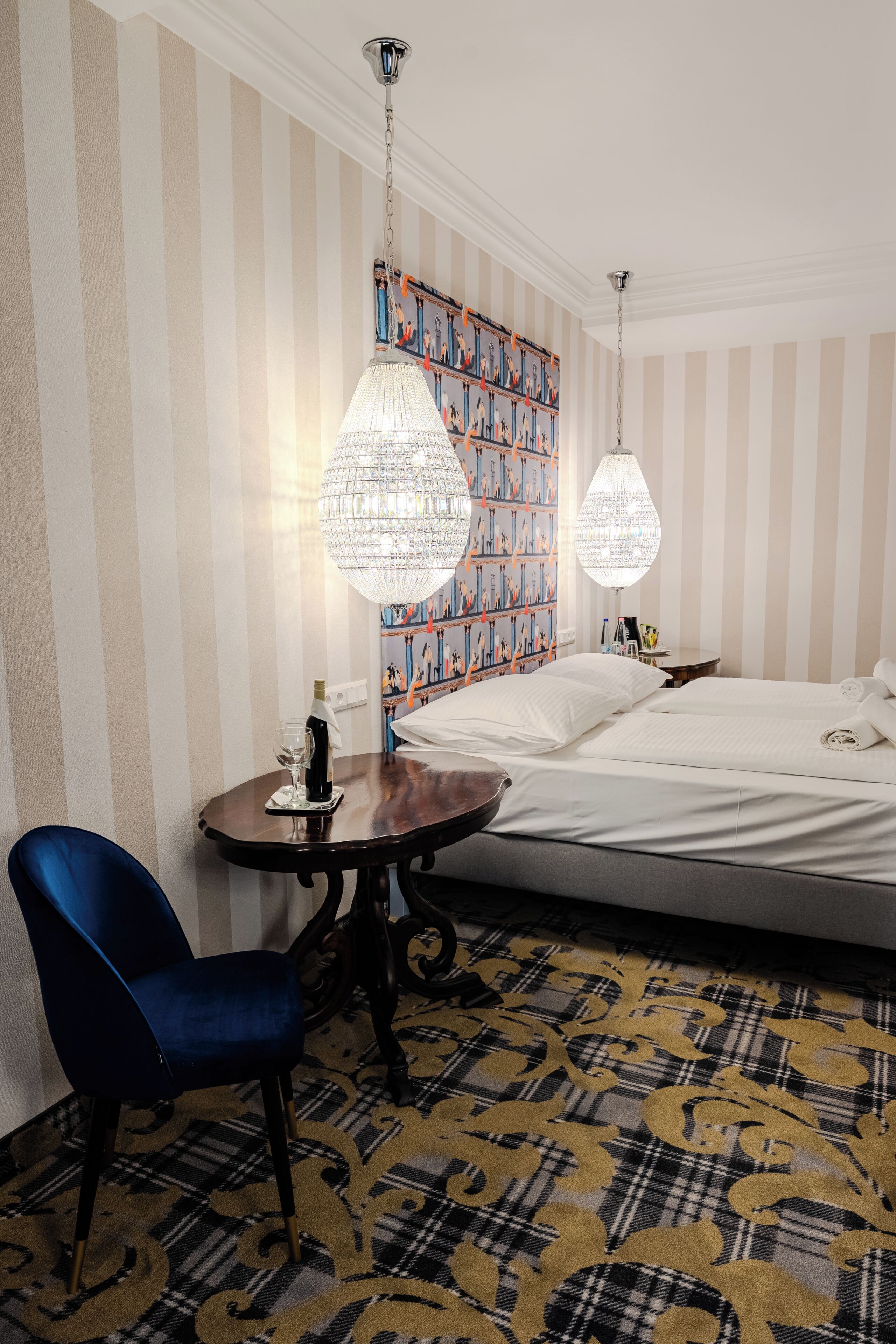Habitación de hotel con cama, escritorio con silla, dos candelabros de cristal colgantes y una alfombra estampada.