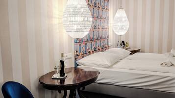 Habitación de hotel con cama, escritorio con silla, dos candelabros de cristal colgantes y una alfombra estampada.