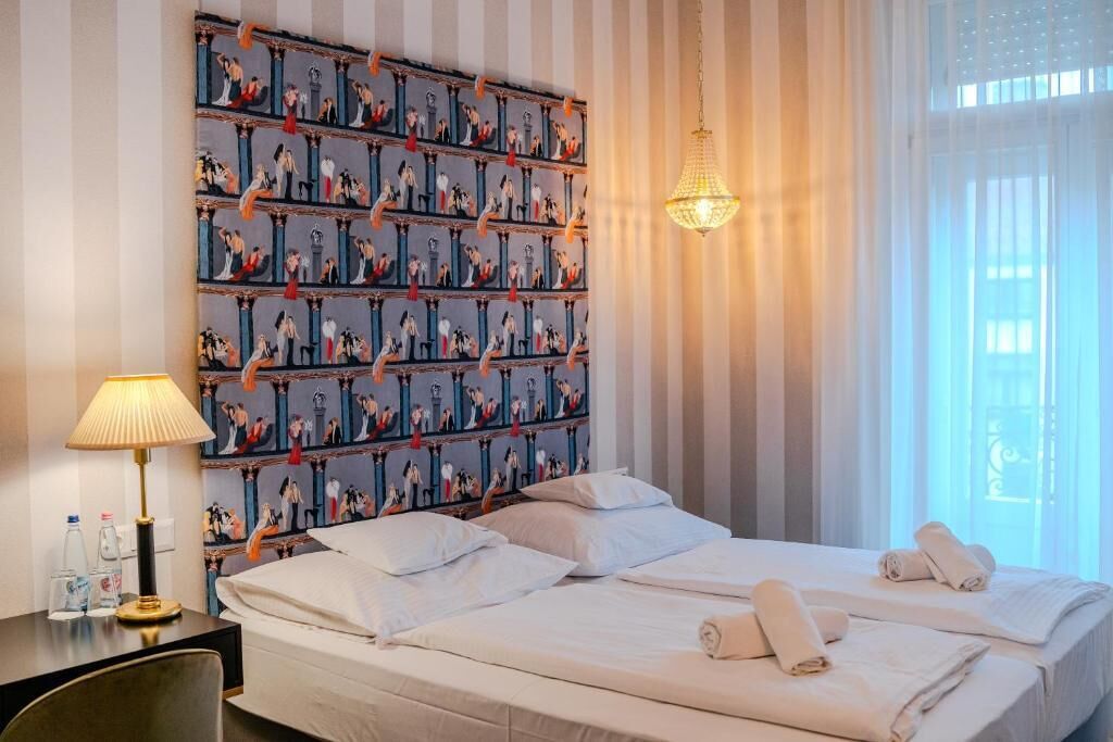 Una habitación de hotel con una cama, almohadas, una mesita de noche con lámpara y una pared con un diseño repetido.