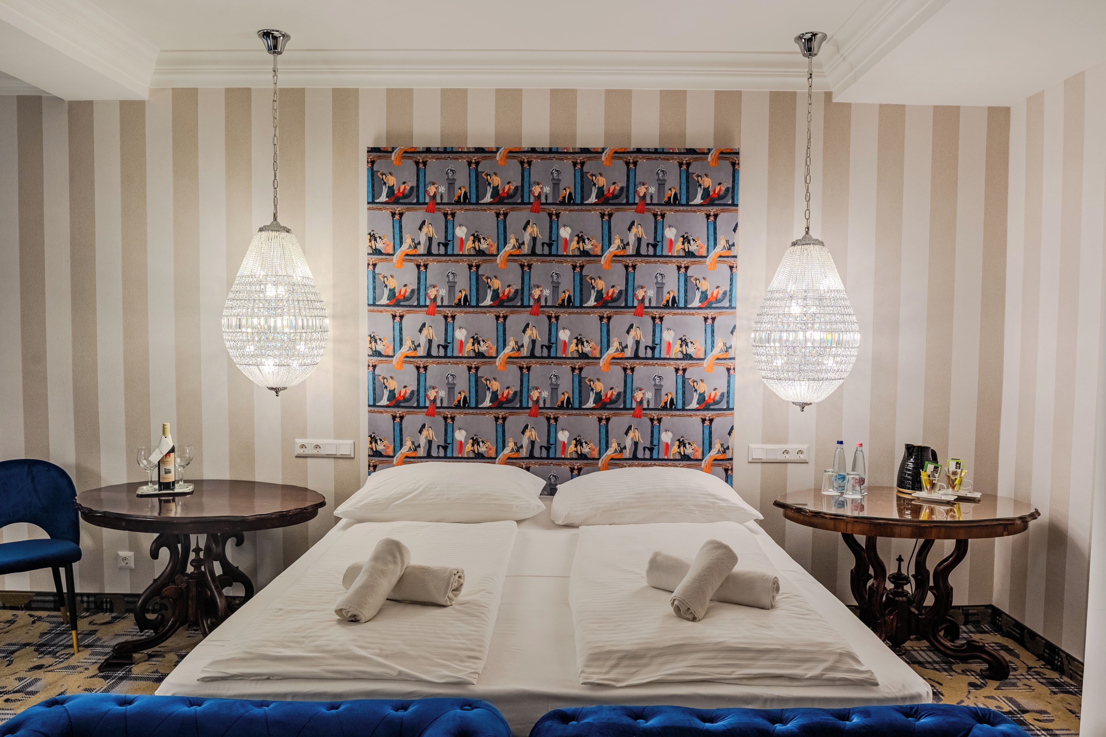 A hotel room with a large bed, two bedside tables, a chandelier, and a patterned wallpaper.