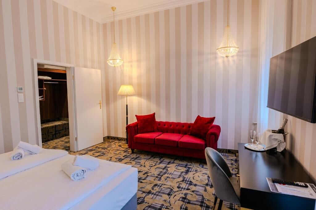 A hotel room with a bed, a red sofa, a desk with a television, and a chandelier.