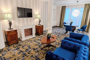 Quarto de hotel com televisão de tela plana, duas luminárias, lareira, tapete estampado, sofá azul e área de refeições com cadeiras e mesa.