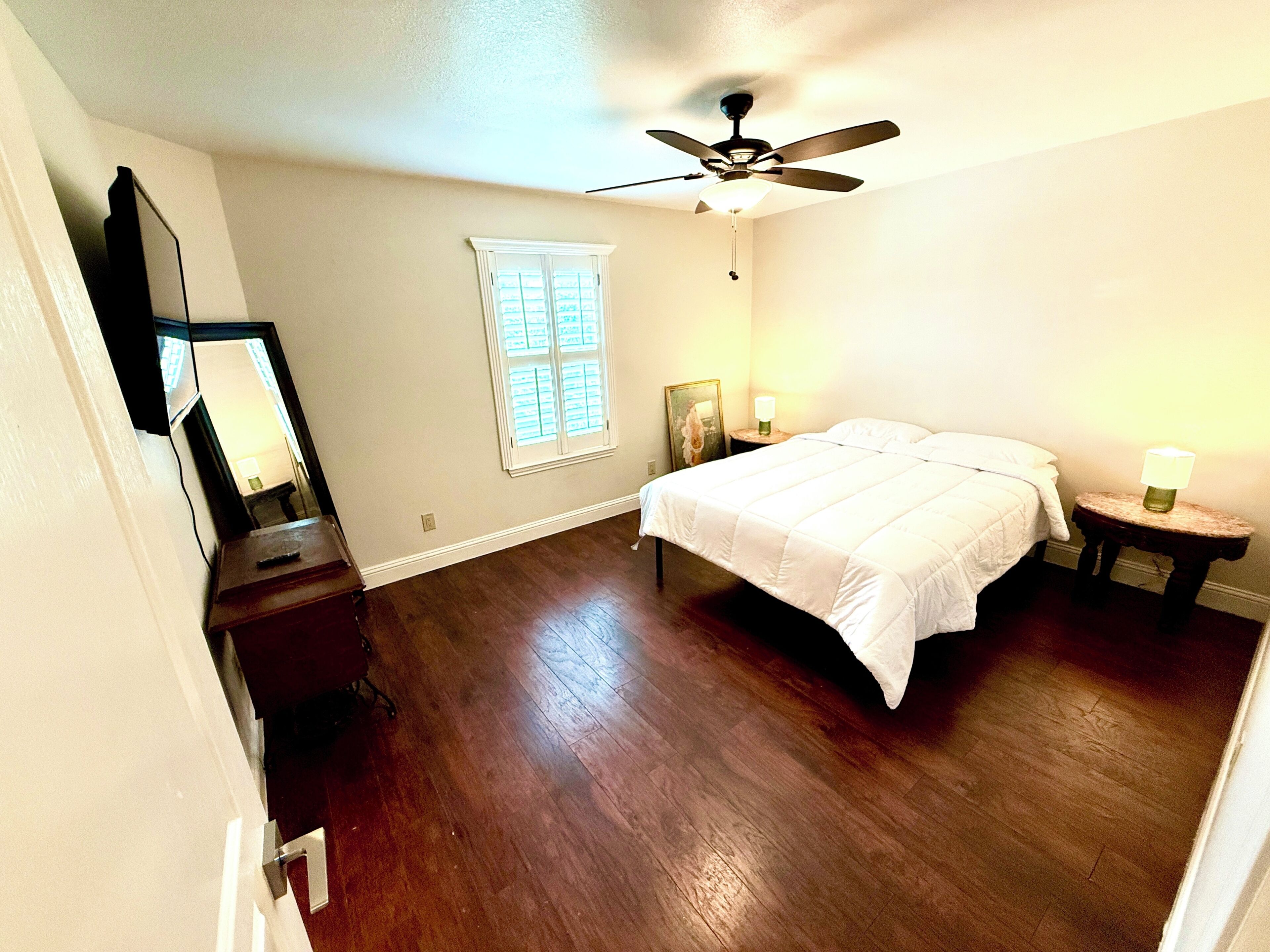 A bedroom with a bed, a ceiling fan, a window with blinds, a nightstand with a lamp, and a dresser.