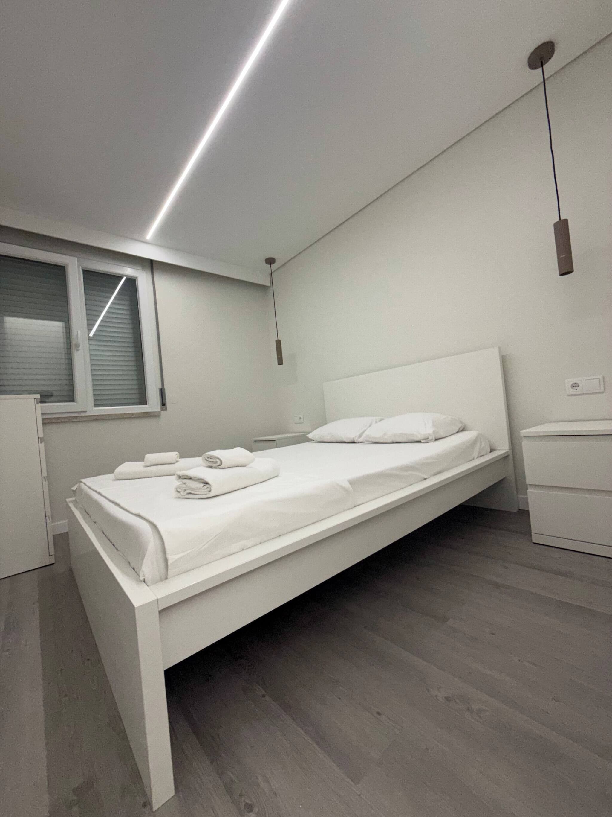 A modern bedroom with a single bed, a bedside table, a nightstand, a window with blinds, and a ceiling light fixture.