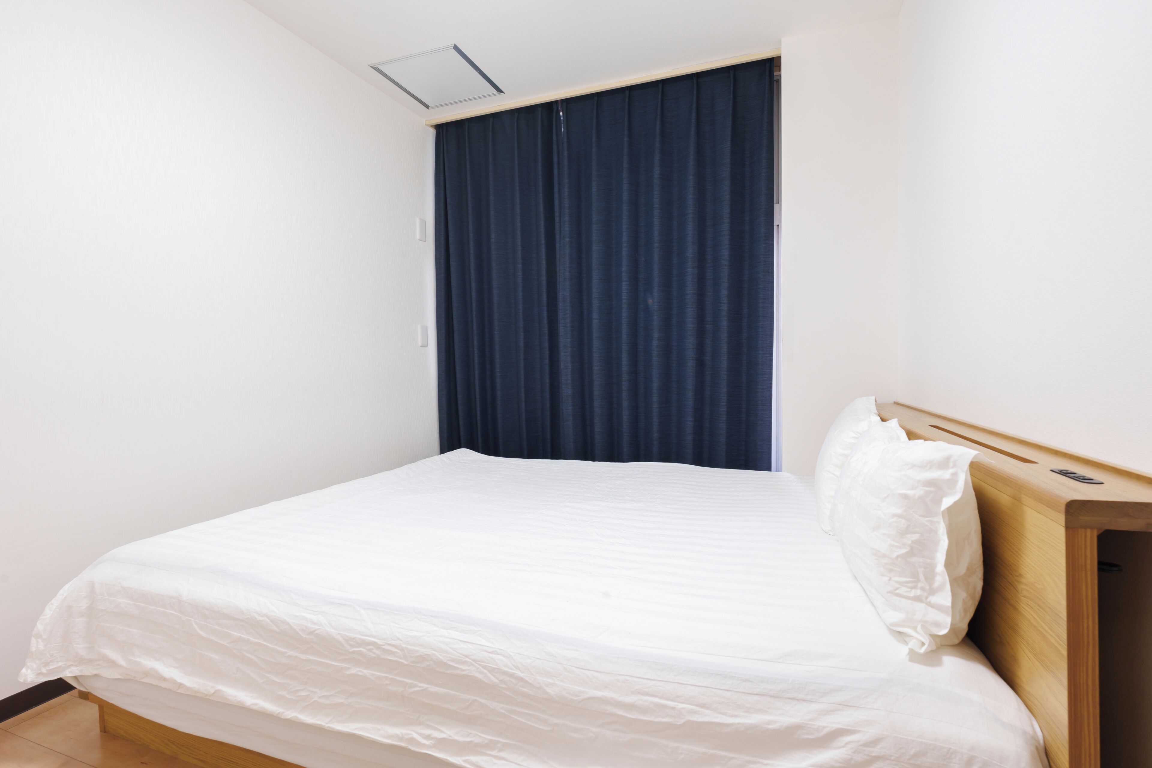 Una cama individual con cabecera de madera en una habitación con paredes blancas y una cortina azul oscuro.