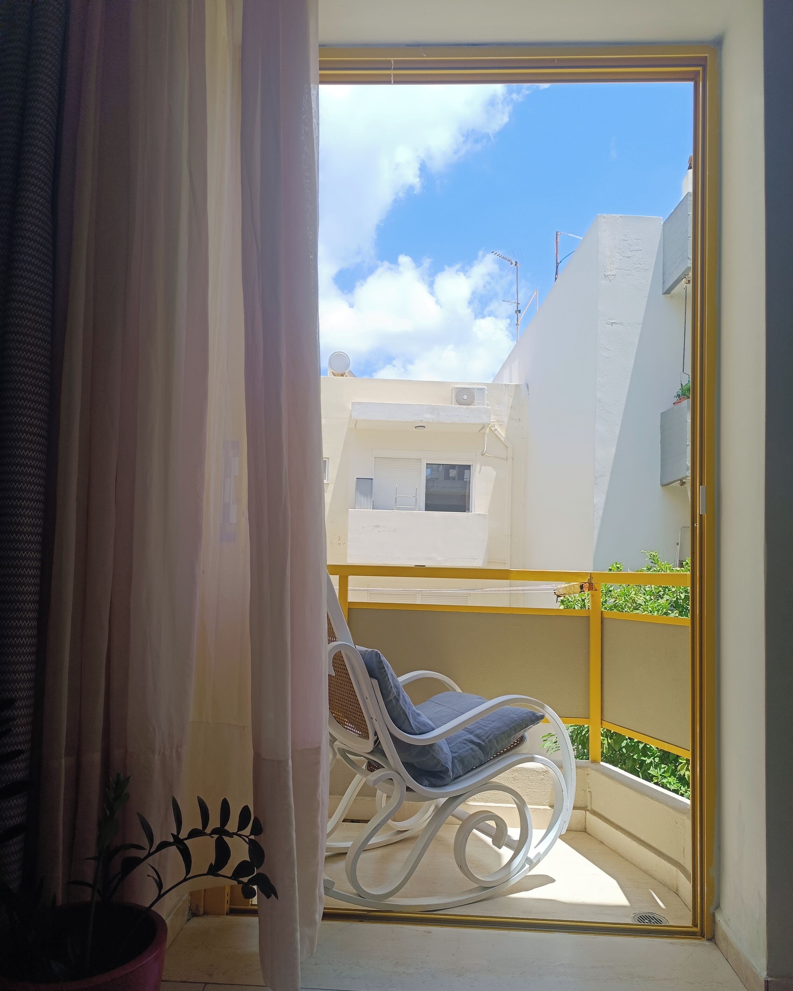 A balcony with a white chair, a potted plant, and a view of a building with a white facade and a yellow railing.