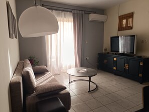 Een woonkamer met een bank, een salontafel, een televisie op een kast, en een raam met gordijnen.