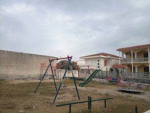 Ein Spielplatz mit Schaukeln und einer Rutsche, ein Gebäude mit rotem Ziegeldach und eine Betonmauer im Hintergrund.
