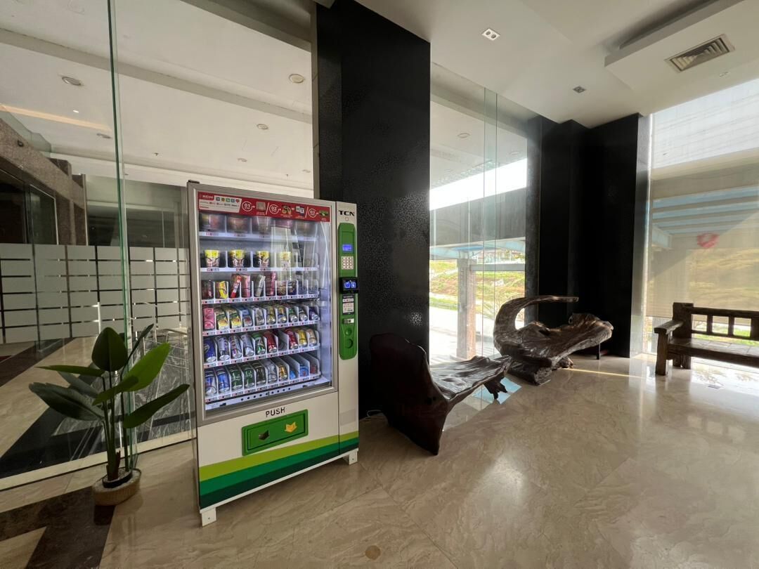 A vending machine with snacks and a push button, a bench, and a potted plant in a modern lobby with glass walls and a view outside.