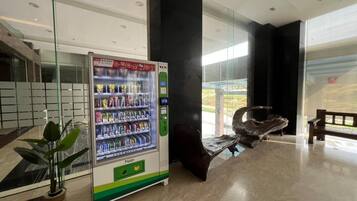 A vending machine with snacks and a push button, a bench, and a potted plant in a modern lobby with glass walls and a view outside.