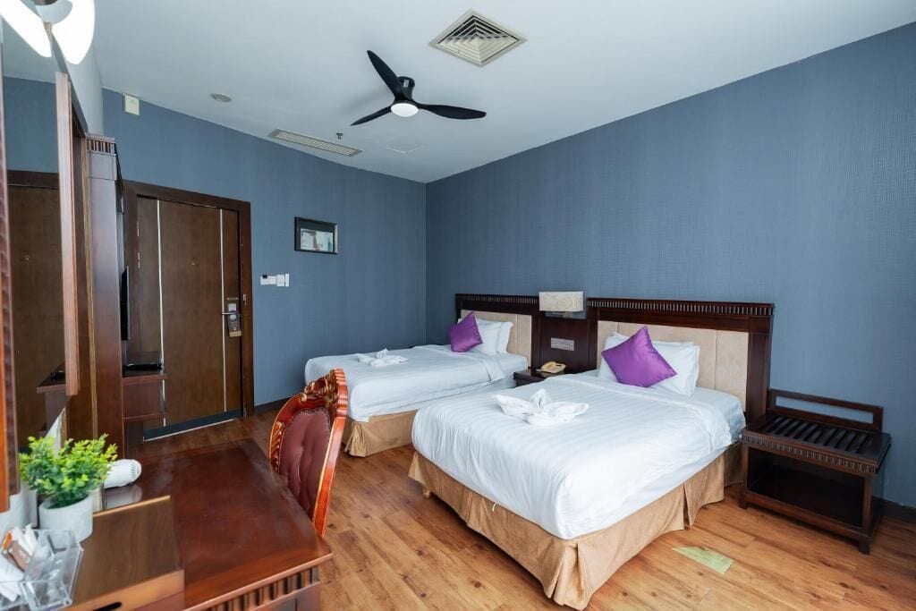 A hotel room with two beds, a desk, a chair, a ceiling fan, and a small table.