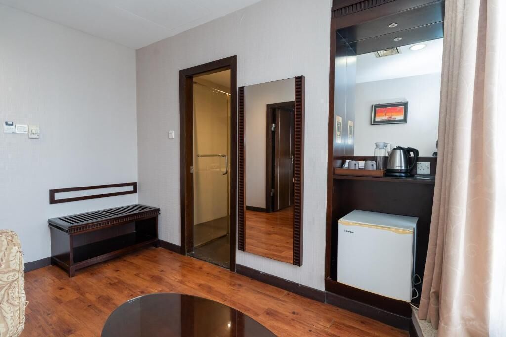 A hotel room with a wooden bench, a small refrigerator, a mirror, and a door leading to a bathroom.