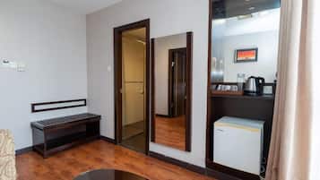 A hotel room with a wooden bench, a small refrigerator, a mirror, and a door leading to a bathroom.