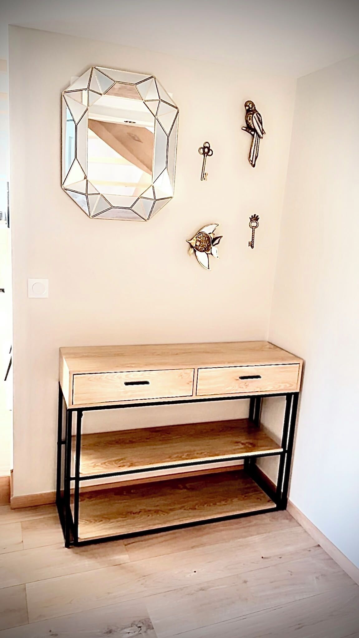 Un bureau en bois doté de tiroirs et d’une étagère, un miroir aux motifs géométriques et des clés décoratives accrochées au mur.