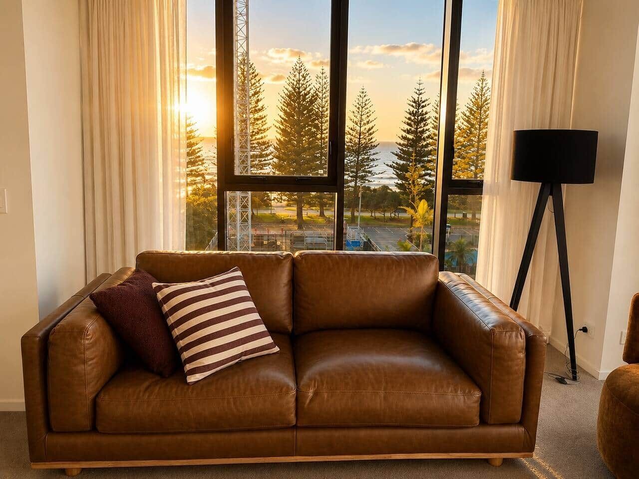 A brown leather sofa with two pillows, a black floor lamp, and a view of trees and a building through large windows.