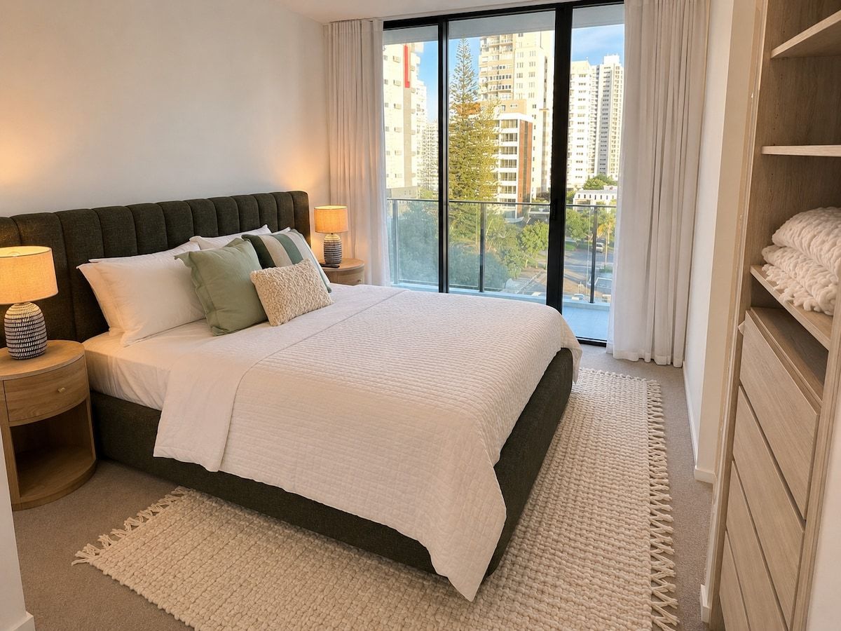 A hotel room with a large bed, a nightstand, a lamp, a view of the city through a balcony, and a wardrobe.