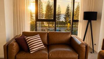 A brown leather sofa with two pillows, a black floor lamp, and a view of trees and a building through large windows.