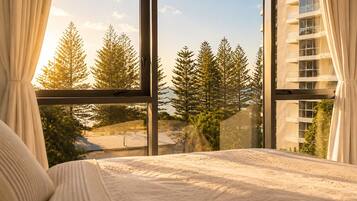 A bedroom with a large window, a bed with white bedding, and a view of trees and buildings.