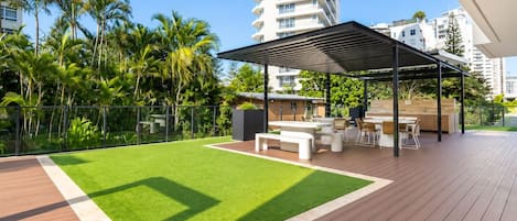 A modern outdoor patio with a wooden deck, a grassy area, and a covered seating area with tables and chairs.