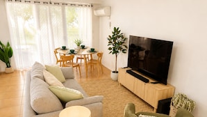 Ein Wohnzimmer mit einer Couch, einem Fernseher auf einem Holzschrank, einem Esstisch mit Stühlen und einer Topfpflanze.