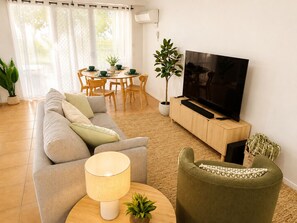 Ein Wohnzimmer mit einer Couch, einem Fernseher auf einem Holzschrank, einem Esstisch mit Stühlen und einer Topfpflanze.