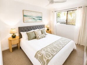 Ein ordentlich eingerichtetes Schlafzimmer mit einem Bett, zwei Nachttischlampen, einem Deckenventilator, einem Fenster mit Vorhängen und einem an der Wand hängenden Gemälde.