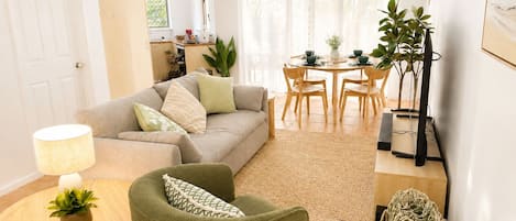 Uma sala de estar com sofá, poltrona e uma mesa de jantar com cadeiras. Há uma luminária em uma mesa lateral e uma planta no chão.