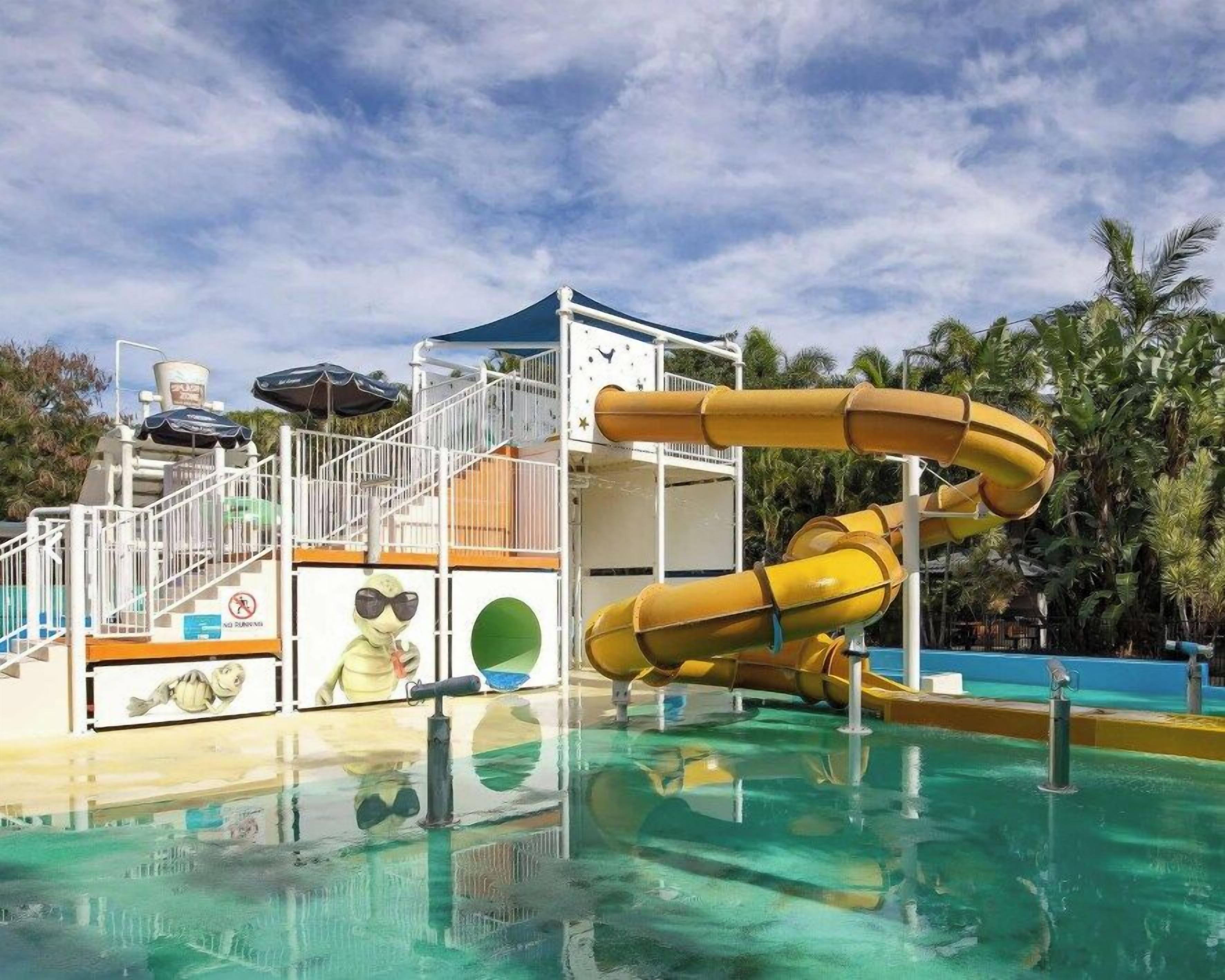 A water park with a large yellow slide, a pool, and a covered area with stairs.