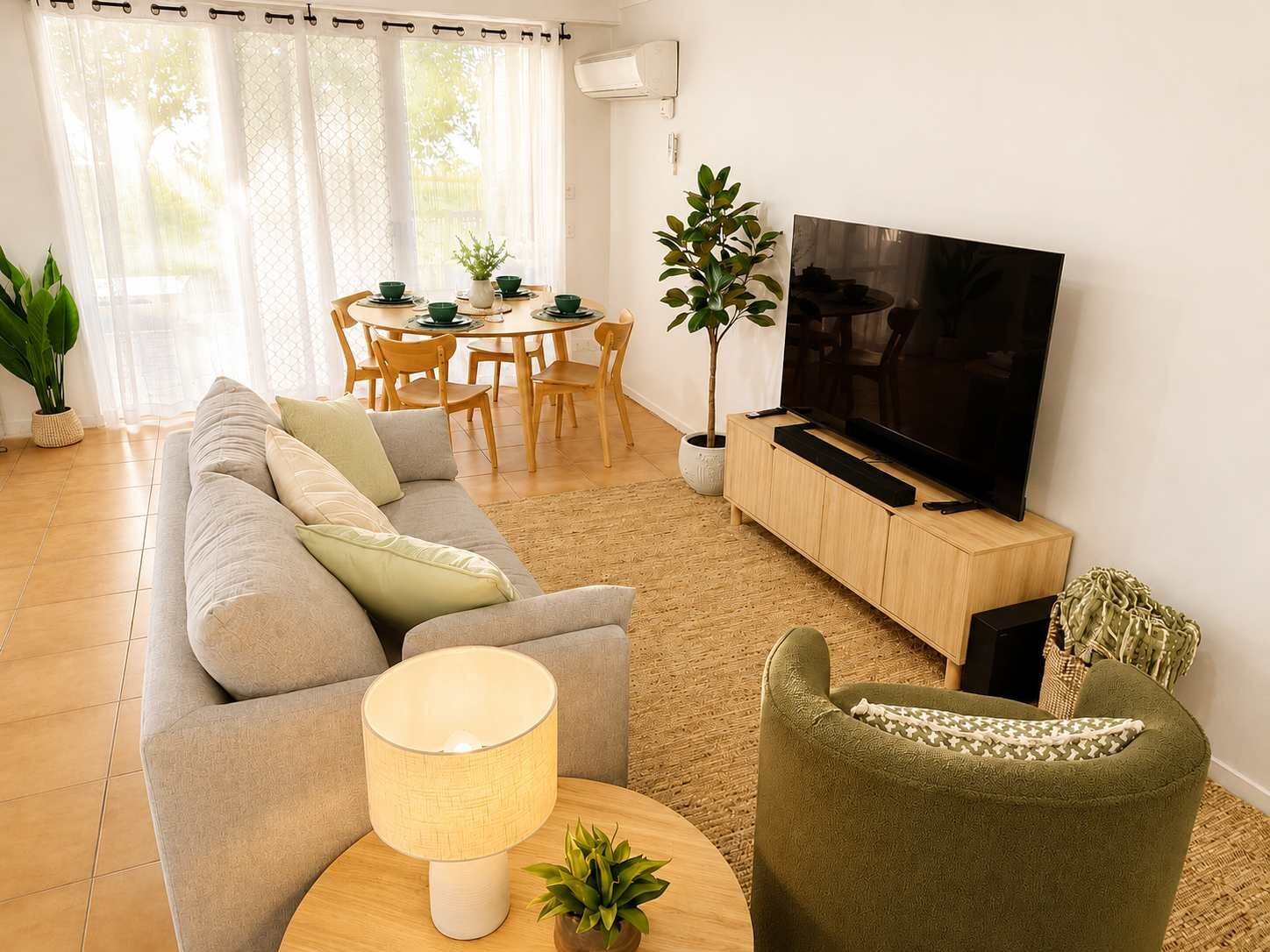 A living room with a sofa, a green armchair, a TV on a wooden stand, a dining table with chairs, and a potted plant.