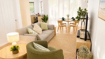 A living room with a sofa, armchair, and a dining table with chairs. There is a lamp on a side table, a potted plant, and a television on a stand.