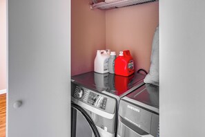 Un área de lavandería con una lavadora y secadora Samsung SuperSpeed Steam SmartTop, una botella de detergente blanco, una botella de detergente gris y una botella de detergente rojo.