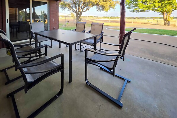 Un patio exterior con una mesa y sillas, un vidrio corredizo que da a un área techada y vistas a un campo cercado.