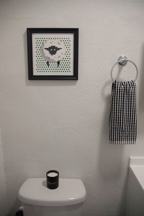 A bathroom with a white toilet, a framed pixel art of a sheep, and a black and white checkered towel hanging on a metal ring.
